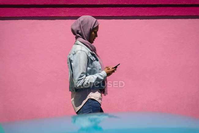 Vista lateral da bela mulher de raça mista usando telefone celular enquanto caminha do beco em um dia ensolarado — Fotografia de Stock