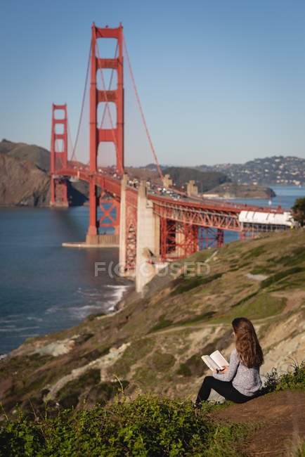 Вид ззаду красива жінка читає книгу, сидячи на схилі гори — стокове фото