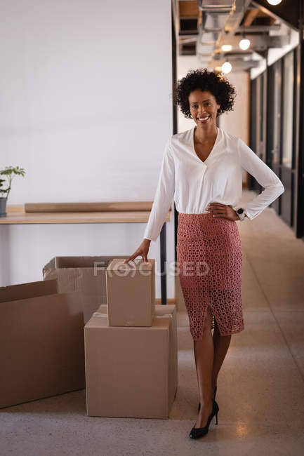 Porträt einer schönen gemischtrassigen Geschäftsfrau, die mit der Hand auf der Hüfte im Büro steht und die andere auf einem Karton. sie blickt und lächelt in die Kamera — Stockfoto