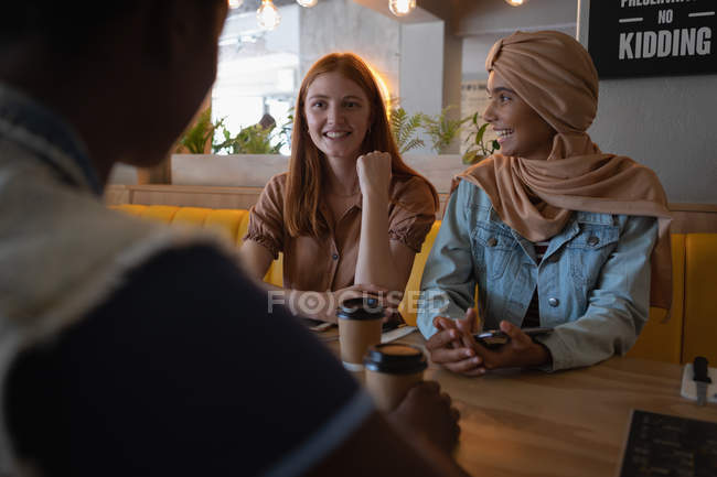 Vista frontal de amigos femeninos de raza mixta interactuando entre sí mientras toman café en el restaurante - foto de stock