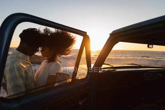 Seitenansicht eines romantischen afrikanisch-amerikanischen Paares, das bei Sonnenuntergang am Strand in der Nähe des Autos steht — Stockfoto