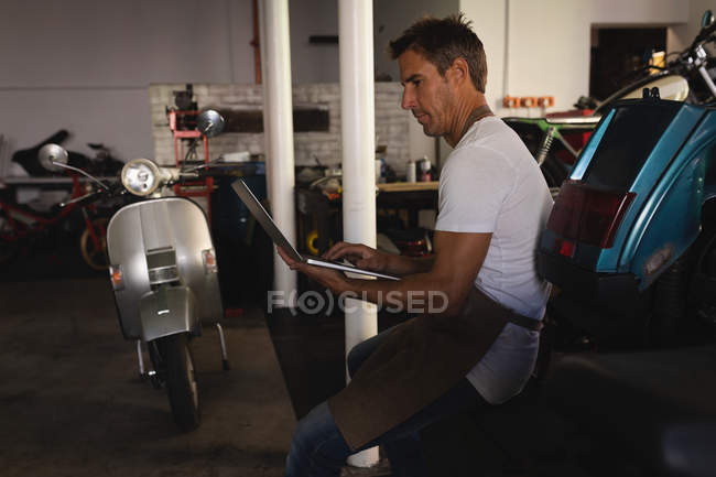 Вид сбоку на кавказского мотомеханика с помощью ноутбука, сидя в гараже — стоковое фото