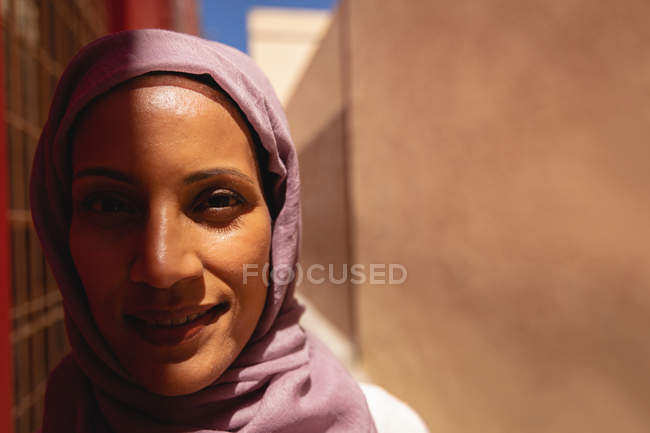 Primer plano de la mujer de raza mixta sonriendo y mirando a la cámara mientras está de pie en el callejón en un día soleado - foto de stock