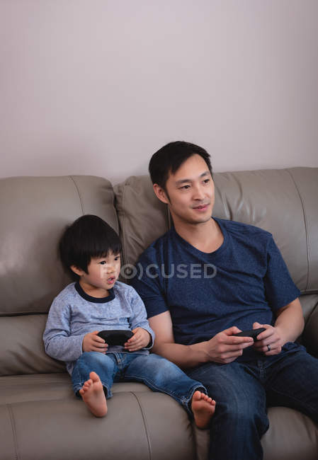 Vista frontal de feliz padre asiático e hijo jugando juntos en los videojuegos mientras están sentados en el sofá en casa - foto de stock