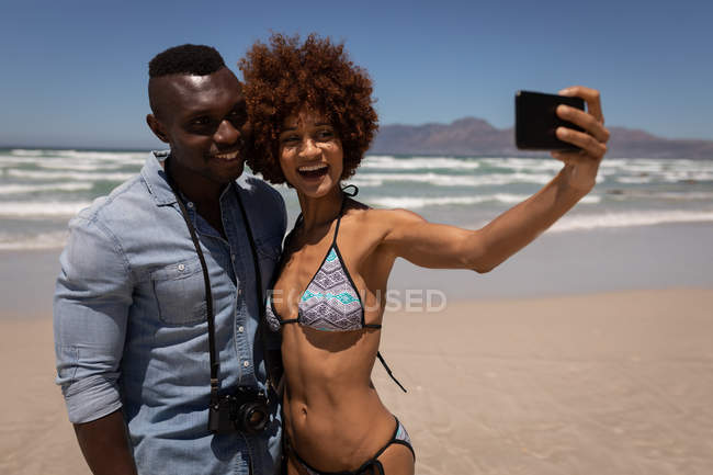 Vista frontal de una linda pareja multiétnica tomando selfie con teléfono móvil en la playa - foto de stock