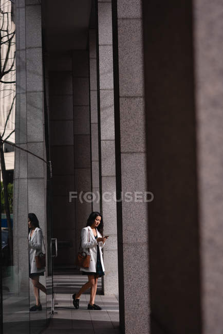 Vue de face de la femme asiatique utilisant un téléphone portable tout en marchant dans le couloir — Photo de stock