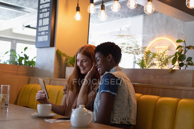 Vista lateral de felices amigas de raza mixta escuchando música con auriculares mientras están sentadas en el restaurante - foto de stock