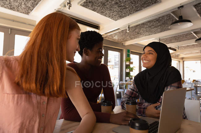 Вид збоку на молоду змішану расу друзі-жінки взаємодіють один з одним, використовуючи ноутбук у кафе — стокове фото