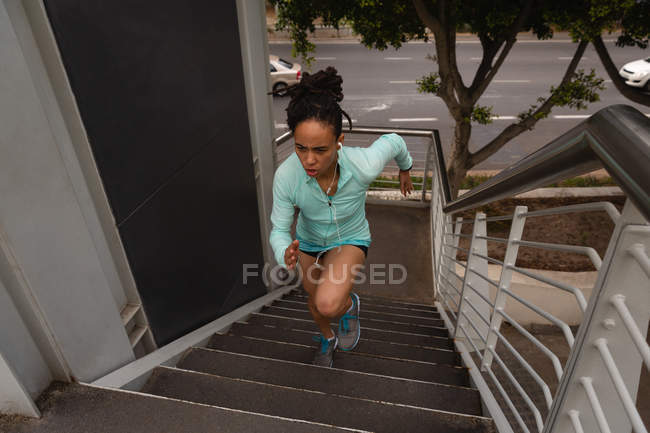 Vista de ángulo alto de la joven mujer de raza mixta escuchando música en los auriculares mientras hace ejercicio en las escaleras de la ciudad - foto de stock