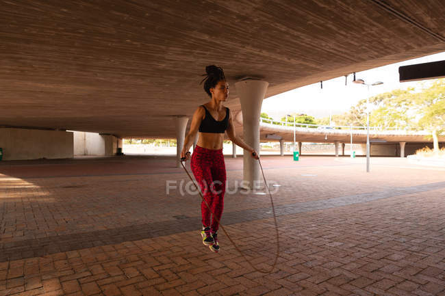Vue de face de la jeune femme de course mixte faisant de l'exercice avec corde à sauter sous un pont dans la ville — Photo de stock