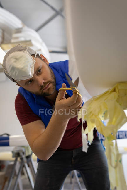 Low angle view of concentrated Caucasian man making a surfboard in surf shop. He is cutting protective veil in workshop — Stock Photo