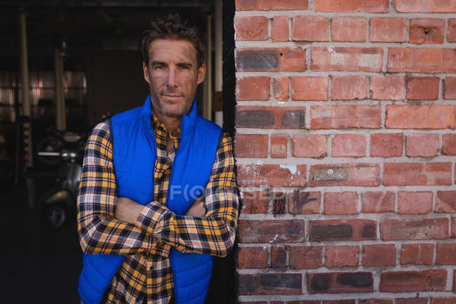 Portrait of Caucasian bike mechanic leaning at brick wall while arms crossed in garage — Stock Photo