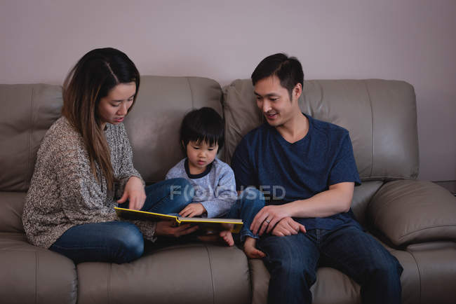 Vista frontal do livro de leitura da família asiática feliz enquanto sentados juntos no sofá em casa — Fotografia de Stock