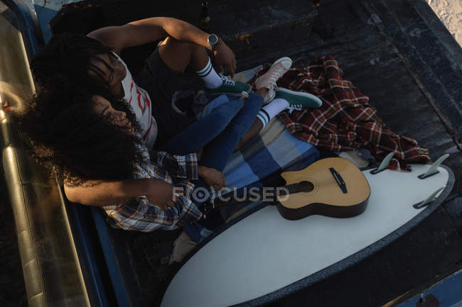 Vista de ángulo alto de la joven pareja afroamericana sentada con el brazo alrededor cerca de la guitarra en el coche en la playa en el día soleado - foto de stock