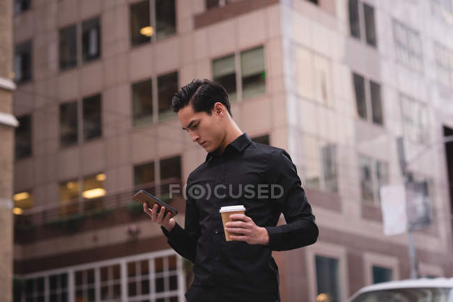 Vue de face du bel homme asiatique utilisant un téléphone mobile dans la rue tout en prenant un café — Photo de stock