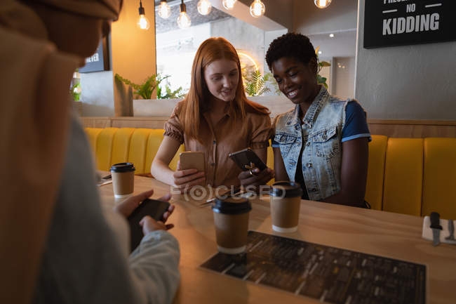 Vista frontal de mestiços amigos do sexo feminino usando telefone celular enquanto sentado no restaurante — Fotografia de Stock
