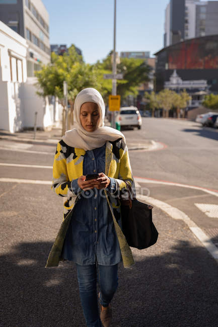 Frontansicht einer Mischlingsfrau, die ihr Handy benutzt, während sie an einem sonnigen Tag auf der Straße spaziert — Stockfoto