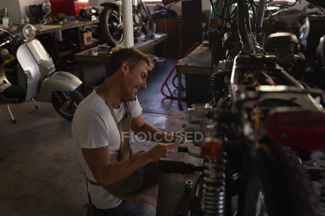 Vista lateral de la bicicleta caucásica mecánico tomando el teléfono móvil durante la reparación de la bicicleta en el garaje - foto de stock