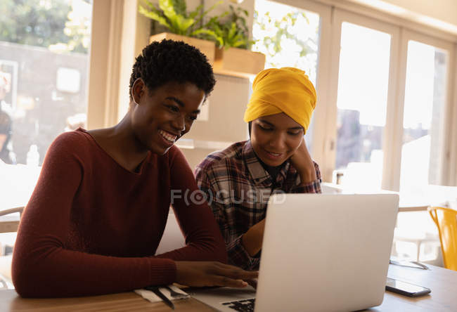 Vista lateral de amigas afroamericanas usando laptop mientras están sentadas en un restaurante - foto de stock
