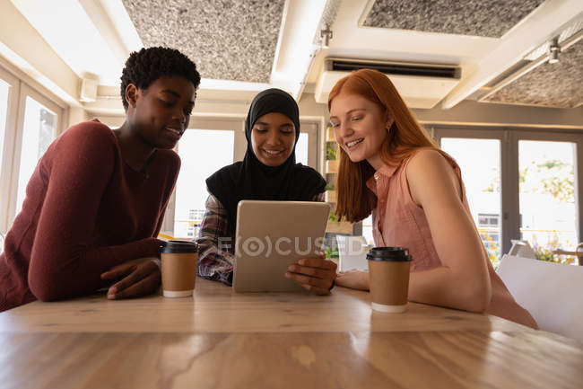 Вид спереду на молоду змішану расу подруги-жінки взаємодіють один з одним, використовуючи цифровий планшет у кафе — стокове фото