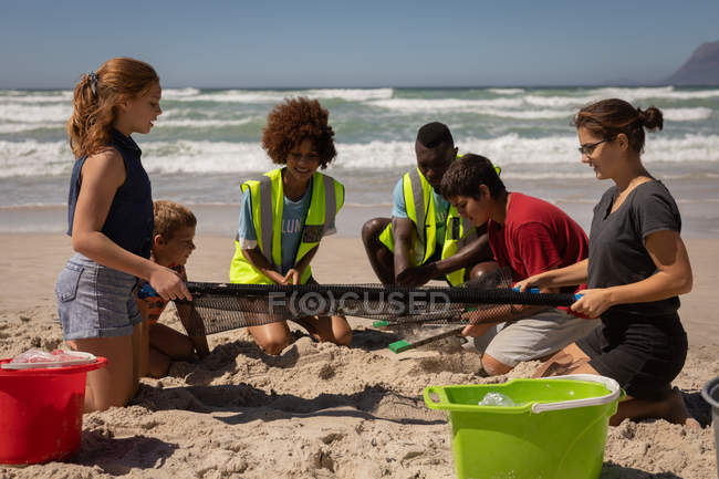 Vue de face d'un groupe de bénévoles multiethniques qui trouvent des déchets avec un filet alors qu'ils sont assis à genoux sur la plage par une journée ensoleillée — Photo de stock