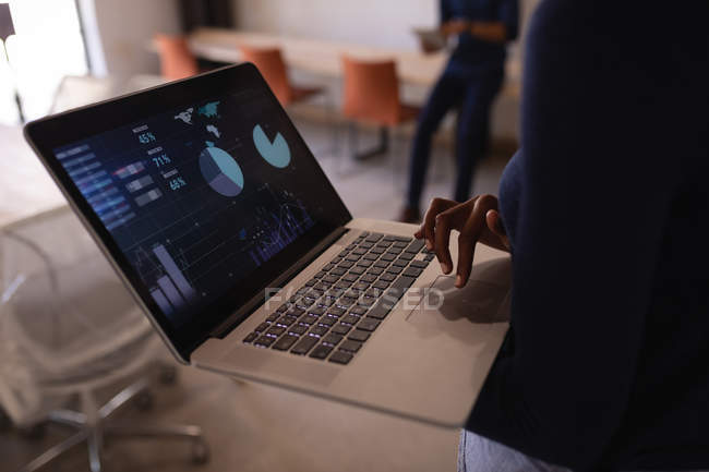 Mittelteil einer Geschäftsfrau mit Laptop, während sie gegen einen Geschäftsmann scrollt, der im Hintergrund auf einem Schreibtisch sitzt — Stockfoto