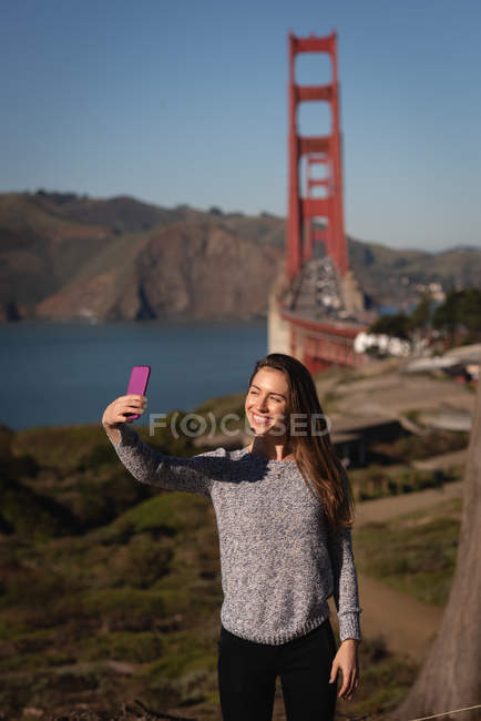 Vista frontal de una hermosa mujer tomando selfie cerca del puente colgante - foto de stock