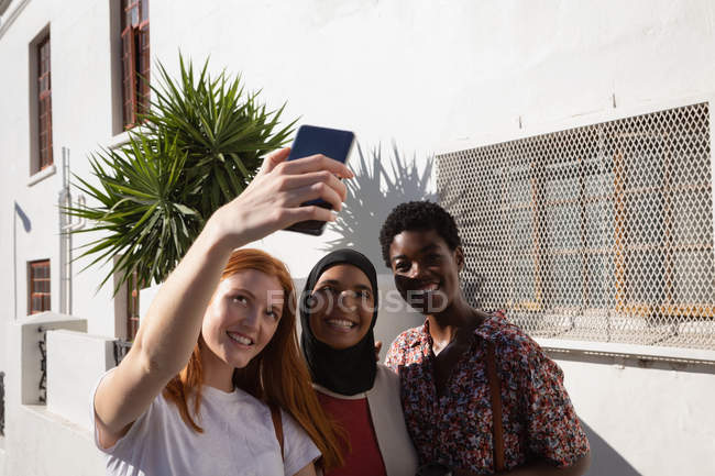 Vista frontal de felices amigas de raza mixta tomando selfie en la calle de la ciudad - foto de stock