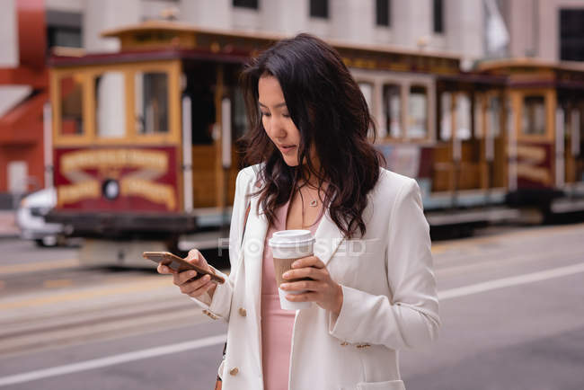 Front view of thoughtful Asian woman using mobile phone while having coffee at street — Stock Photo