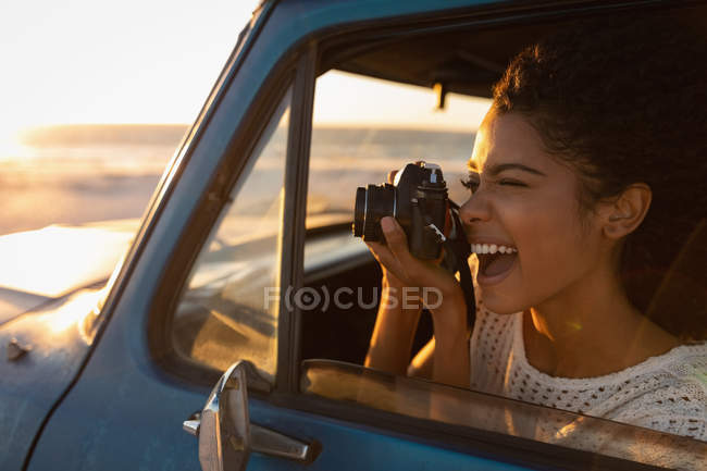 Vista lateral de la hermosa mujer afroamericana feliz tomando fotos con cámara digital mientras está sentado en el coche en la playa al atardecer - foto de stock
