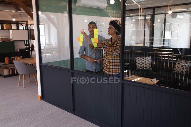 Vista frontal de jovens empresários de raça mista que escrevem sobre notas pegajosas no escritório moderno — Fotografia de Stock