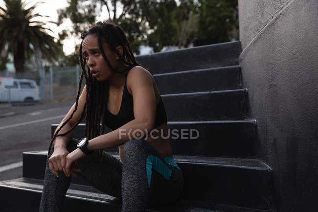 Vista frontale di stanca giovane donna di razza mista rilassante sui gradini della città — Foto stock