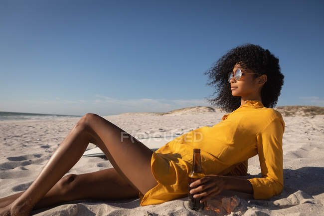 Vista lateral da bela jovem afro-americana que se inclina na praia enquanto toma cerveja no dia ensolarado — Fotografia de Stock