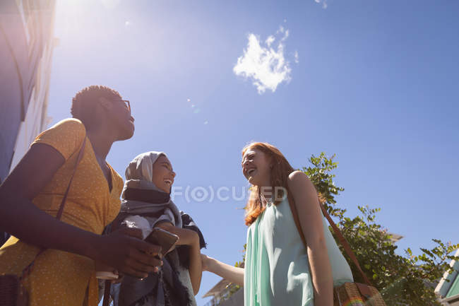 Vista de ángulo bajo de jóvenes amigas de raza mixta que interactúan entre sí en la ciudad en un día soleado - foto de stock
