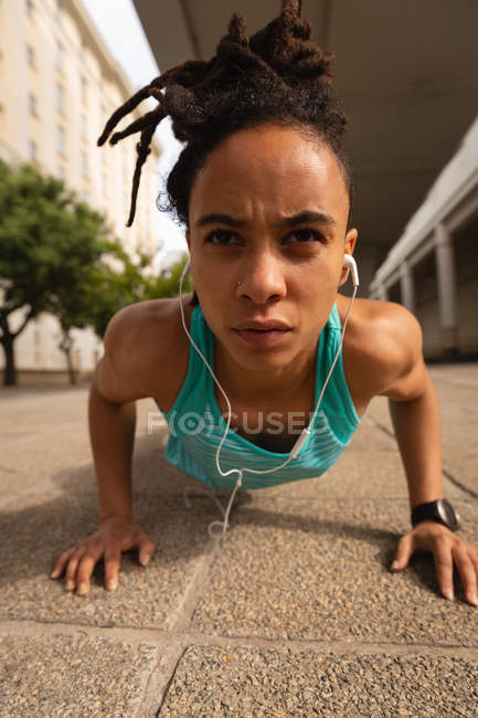 Retrato da jovem mulher de raça mista ouvindo música em fones de ouvido enquanto faz exercício push-up na cidade — Fotografia de Stock
