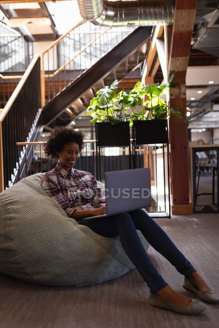 Side view of beautiful Mixed-race businesswoman using laptop while relaxing at bean bag in office against stairs in background — Stock Photo