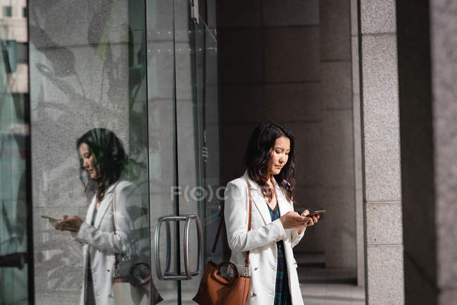 Vue latérale de la femme asiatique utilisant un téléphone portable tout en restant debout dans le couloir — Photo de stock