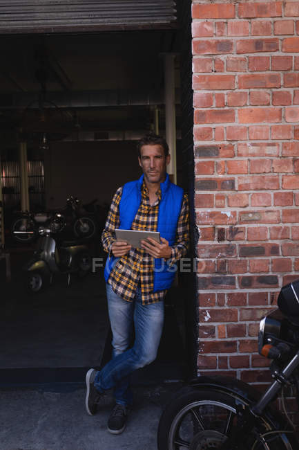 Frontansicht des kaukasischen Fahrradmechanikers lehnt an Ziegelwand, während er digitales Tablet in der Garage hält — Stockfoto
