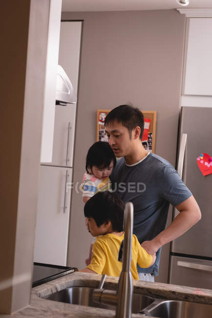 Vorderansicht eines jungen asiatischen Vaters, der seine Kinder hält, während er mit ihnen zu Hause in der Küche spricht — Stockfoto