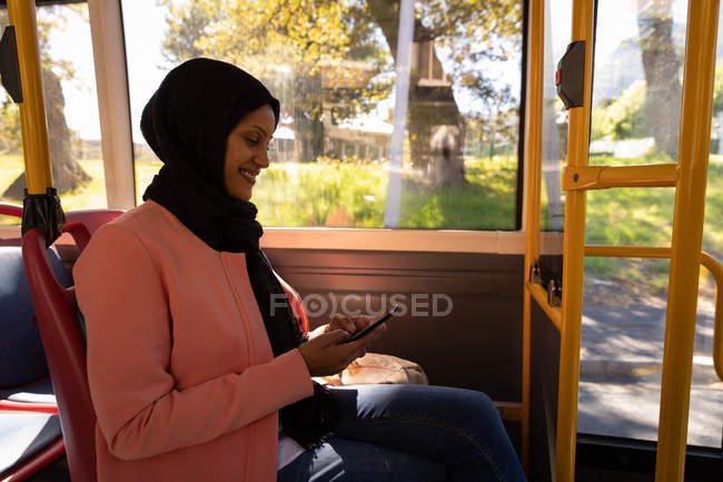 Vista lateral de una hermosa mujer mestiza sonriendo y usando su teléfono móvil mientras viaja en autobús - foto de stock