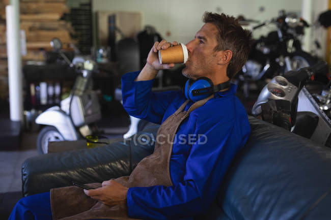 Vista lateral de la bicicleta masculina caucásica mecánico beber café mientras se relaja en sofá azul en el garaje - foto de stock