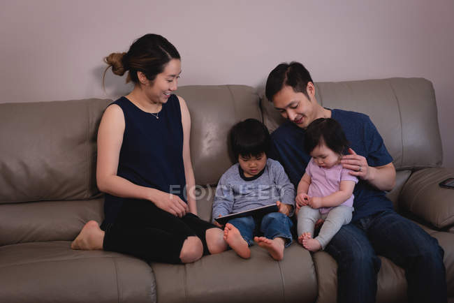 Vista frontal de una feliz familia asiática disfrutando juntos y mirando una tableta digital mientras están sentados en el sofá en casa - foto de stock