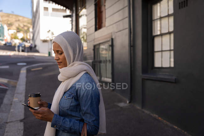 Vista laterale di una donna di razza mista che usa il telefono cellulare e tiene un caffè in piedi sul marciapiede — Foto stock