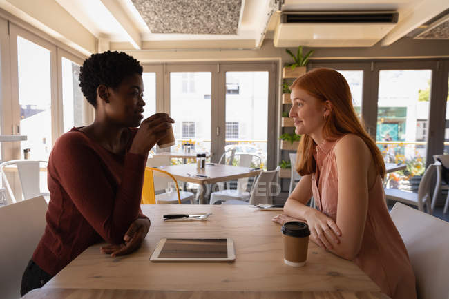Vista lateral de jóvenes amigas de raza mixta interactuando entre sí mientras toman café en un café - foto de stock