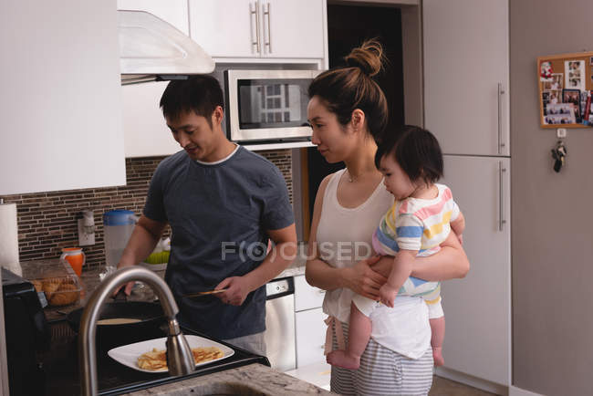 Vista lateral de la madre asiática y el hijo mirando al padre asiático mientras hace panqueques en la cocina en casa - foto de stock