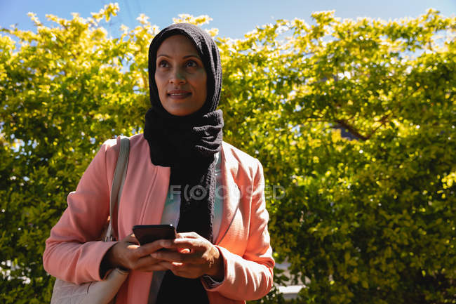 Вид женщины смешанной расы, стоящей с мобильным телефоном на улице в солнечный день — стоковое фото