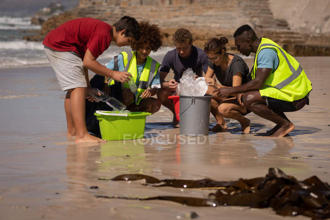 Vista frontal del grupo de jóvenes voluntarios multiétnicos que limpian la playa mientras están sentados en cuclillas frente al cubo en un día soleado - foto de stock