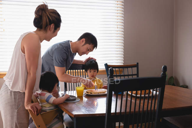 Vista lateral do pai asiático feliz acendendo velas em panquecas enquanto toma café da manhã com sua família na mesa de jantar — Fotografia de Stock