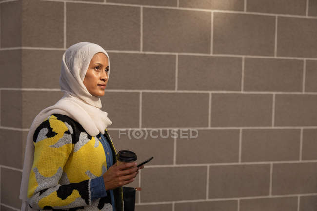 Vista lateral de uma bela mulher de raça mista pensativa em pé com café e telefone celular na frente de uma parede — Fotografia de Stock