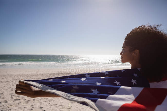 Вид сзади на молодую афроамериканку с американским флагом, стоящую на пляже в солнечный день — стоковое фото
